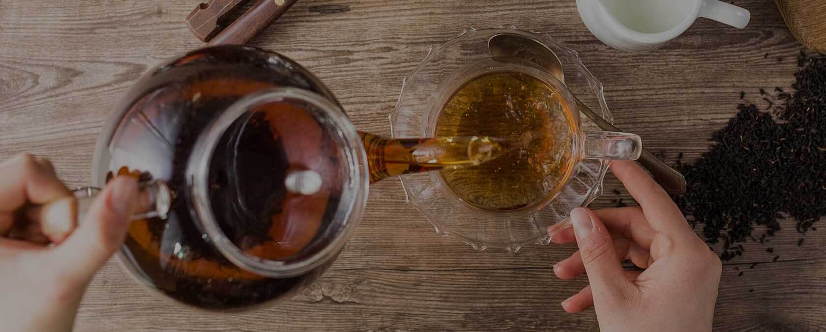 Person gießt Tee aus einer Glasteekanne in eine Tasse; lose Teeblätter und Milchkännchen auf dem Tisch.