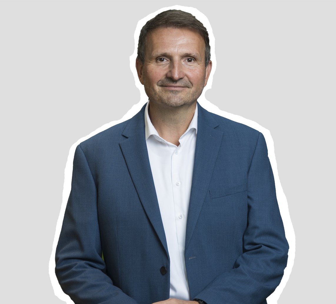 Mann mit kurzen braunen Haaren in blauer Anzugsjacke und weißem Hemd steht vor grauem Hintergrund.