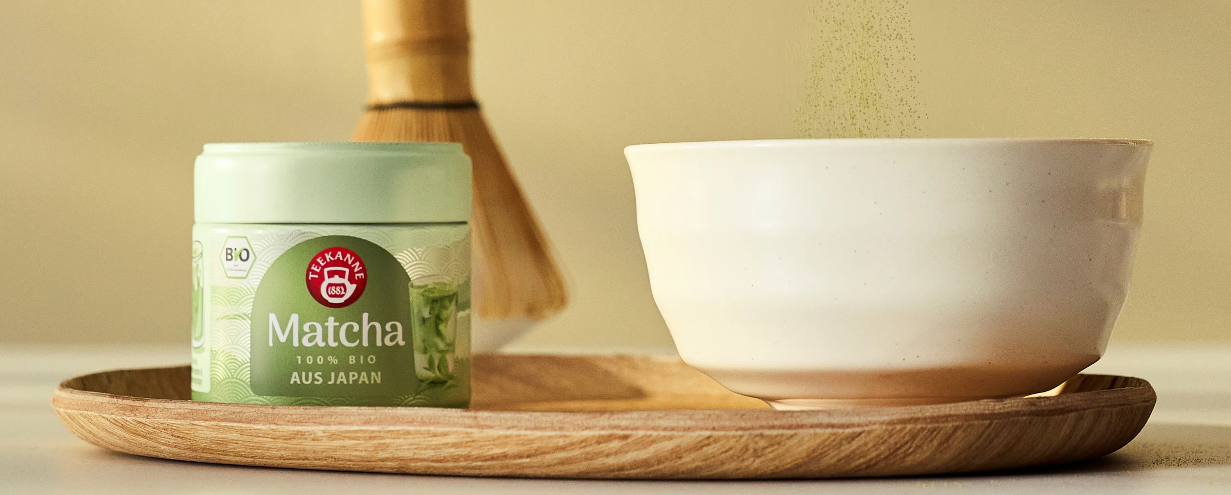 Ein Glas mit japanischem Bio-Matcha auf einem Holztablett, Matcha-Pulver fällt in eine weiße Schale, dahinter ein Bambusbesen.