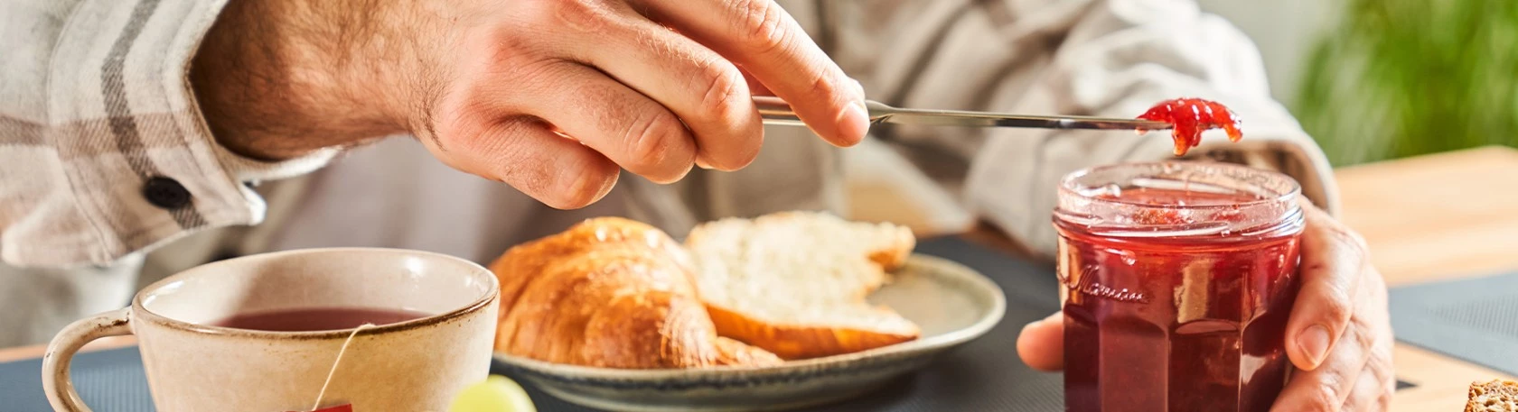 Person streicht rote Marmelade auf Brot; Croissant und Tasse Tee stehen in der Nähe auf dem Frühstückstisch.