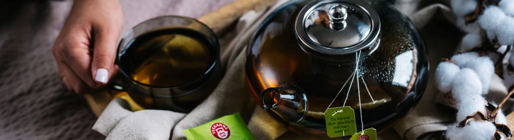 Hand hält eine Glastasse Tee neben Teekanne, Baumwollpflanze und Teeverpackung auf Tablett.