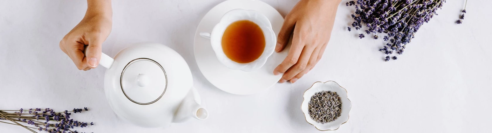 Person gießt Tee aus weißer Teekanne in Teetasse, daneben stehen Lavendelblüten und -knospen.