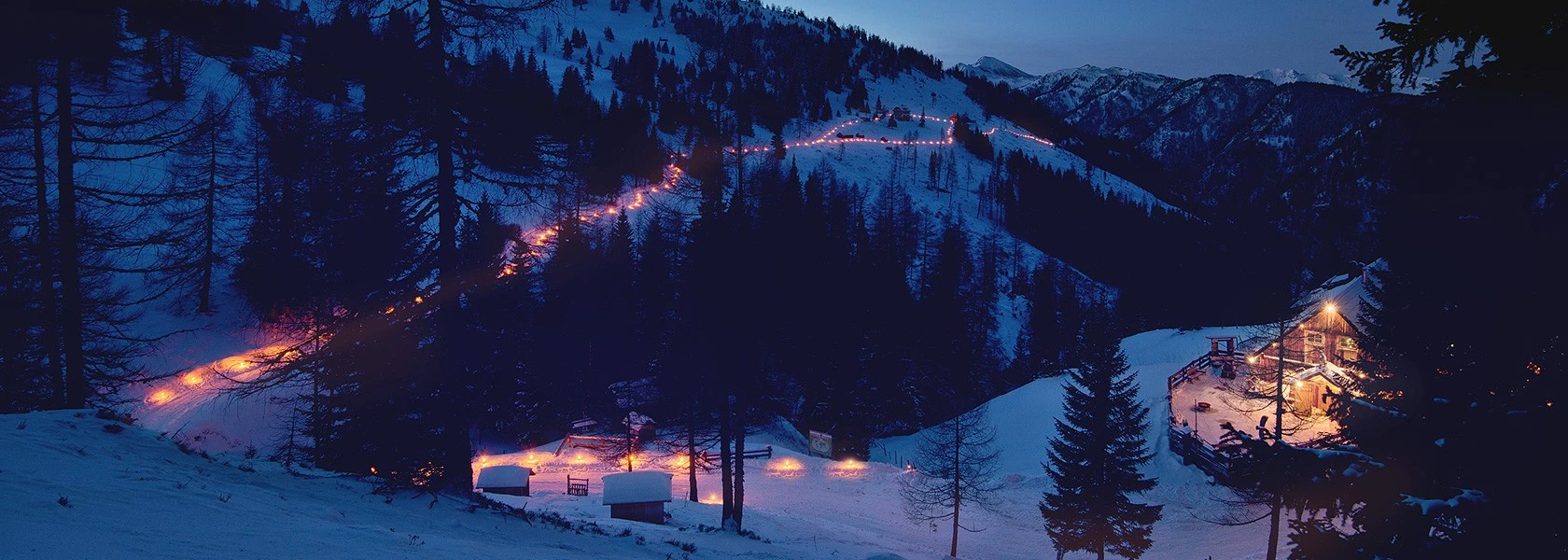 Ein verschneiter Bergpfad mit leuchtenden Lichtern führt zu einer beleuchteten Hütte; im Hintergrund Gipfel und Bäume.