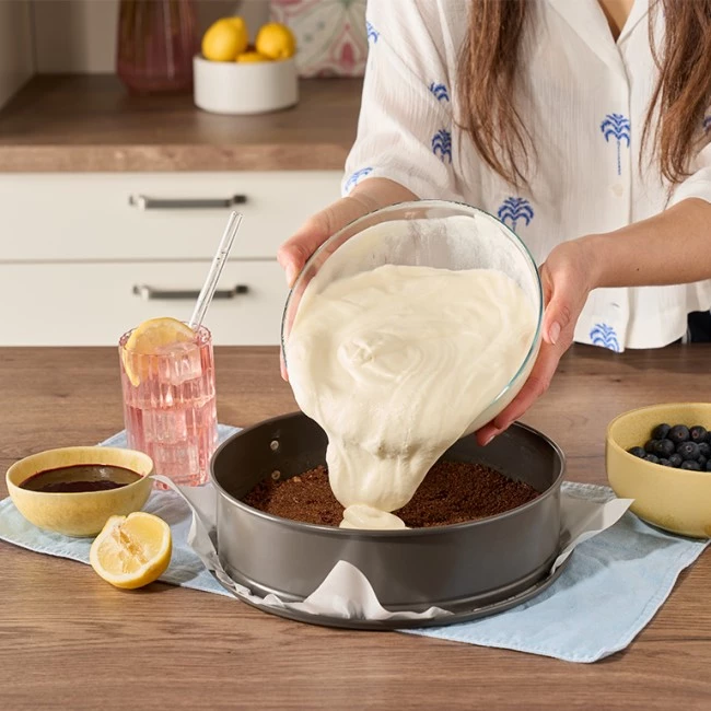 Person gießt Käsekuchenfüllung in Pfanne; daneben stehen rosa Getränk, Schokoladensoße, Zitrone und Blaubeeren.
