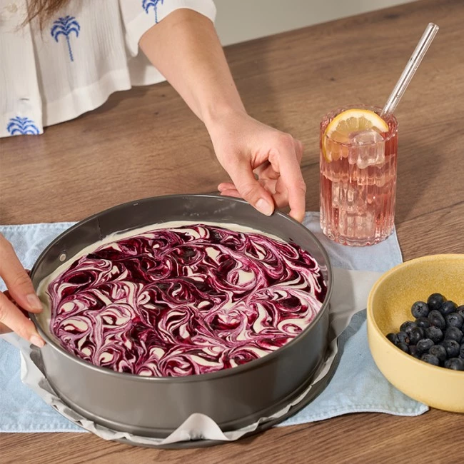 Eine Person stellt einen gewirbelten lila Käsekuchen neben ein rosa Getränk mit Zitrone und eine Schale Blaubeeren.