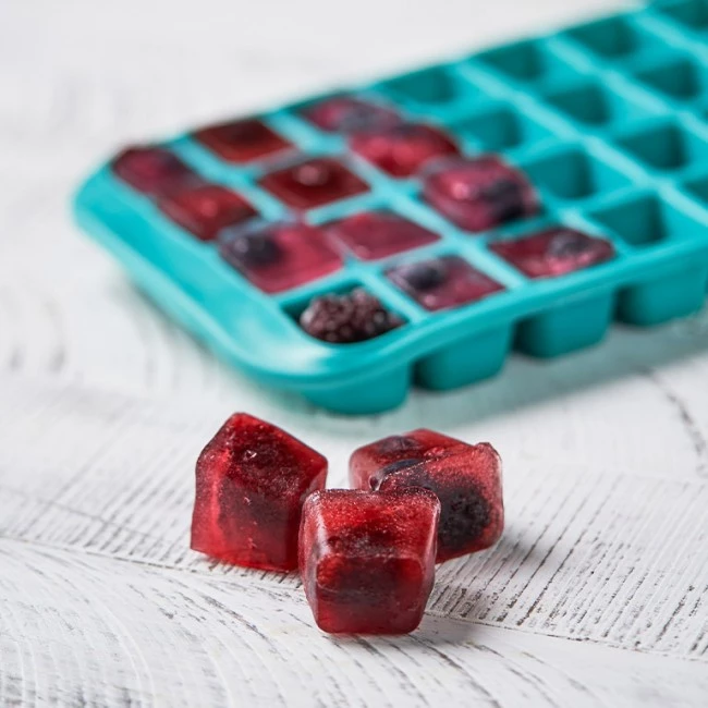 Drei mit Beeren gefüllte rote Eiswürfel vor türkisfarbener Schale auf weißer Holzoberfläche.