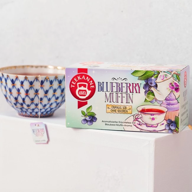 Teekanne Blueberry Muffin-Teedose neben verzierter blau-goldener Teetasse; Tassen und Blaubeeren abgebildet.