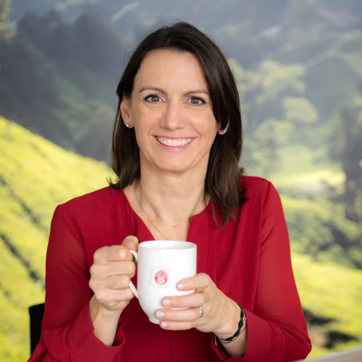 Frau mit braunem Haar, roter Bluse, lächelt, hält weißen Becher, draußen mit viel Grün hinter ihr.