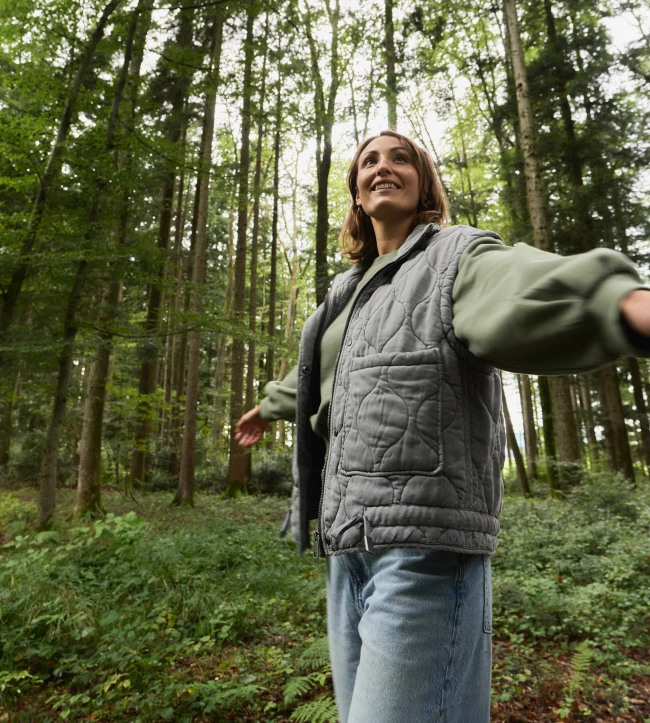 Person in grauer Weste und Jeans, lächelnd, mit ausgestreckten Armen, steht in einem üppigen grünen Wald.