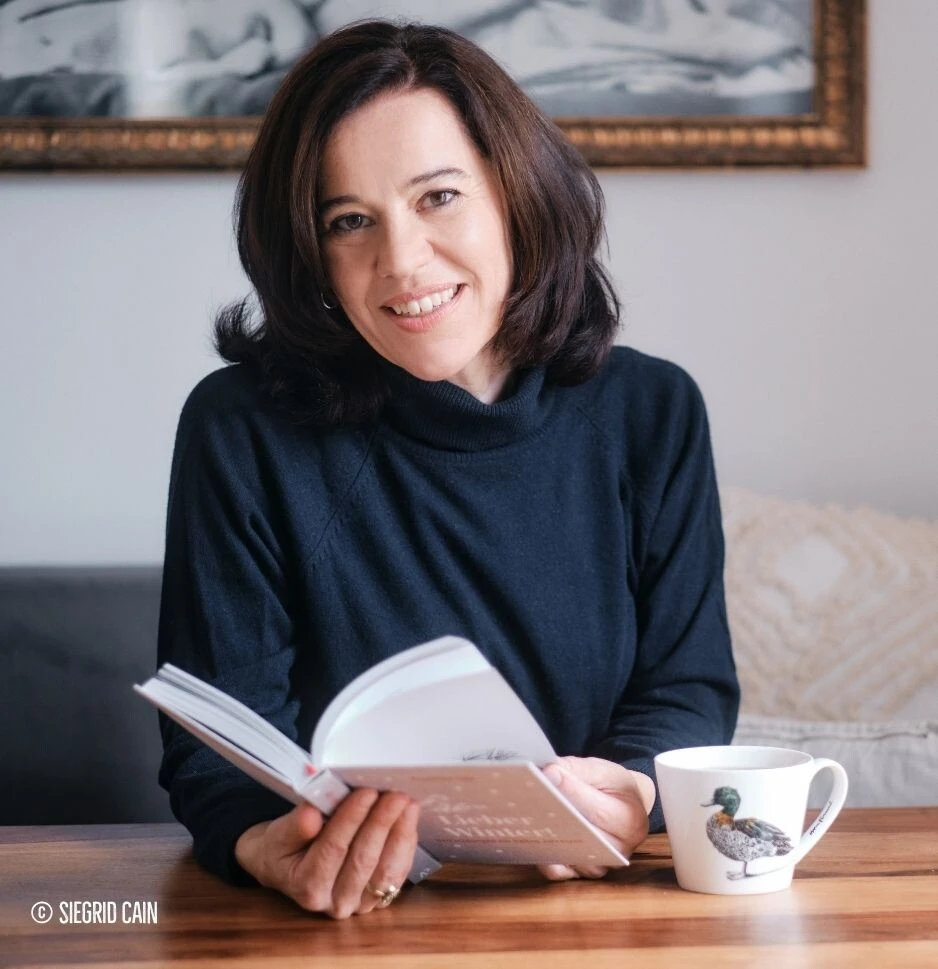 Eine lächelnde Frau mit dunklem Haar liest an einem Holztisch mit einer Tasse und einem gerahmten Bild.