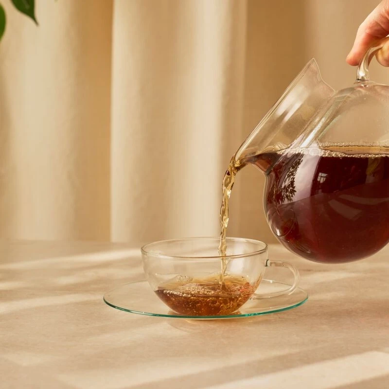 Hand gießt Tee in eine klare Teetasse mit Sonnenlicht und Blattpflanze im Hintergrund.