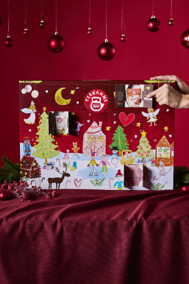 Eine Hand öffnet das Fenster eines festlichen Adventskalenders auf einer roten Tischdecke und gibt den Blick auf eine bunte Teeverpackung im Inneren frei.