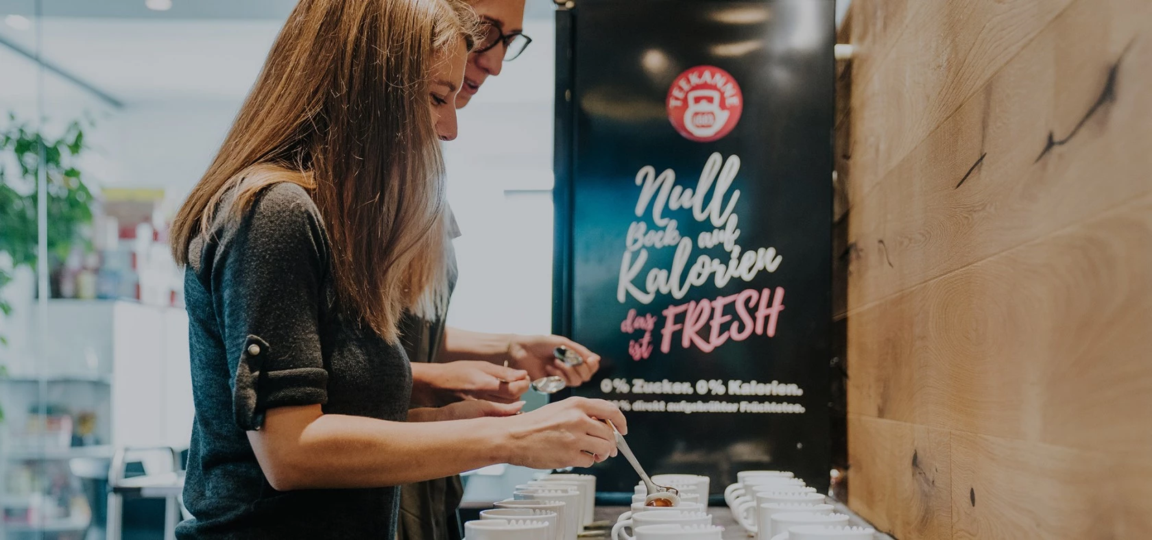 Zwei Personen bereiten an einer Theke Getränke mit weißen Bechern zu, dahinter ein Schild mit der Aufschrift „Kalorienfreies Getränk“.