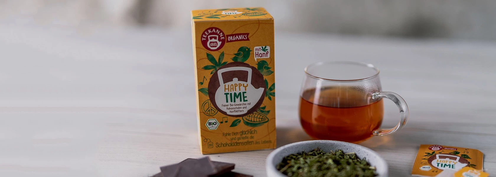 Schachtel Happy Time Hanftee, Tasse aufgebrühten Tee, lose Blätter, Schokolade und einzelner Teebeutel.