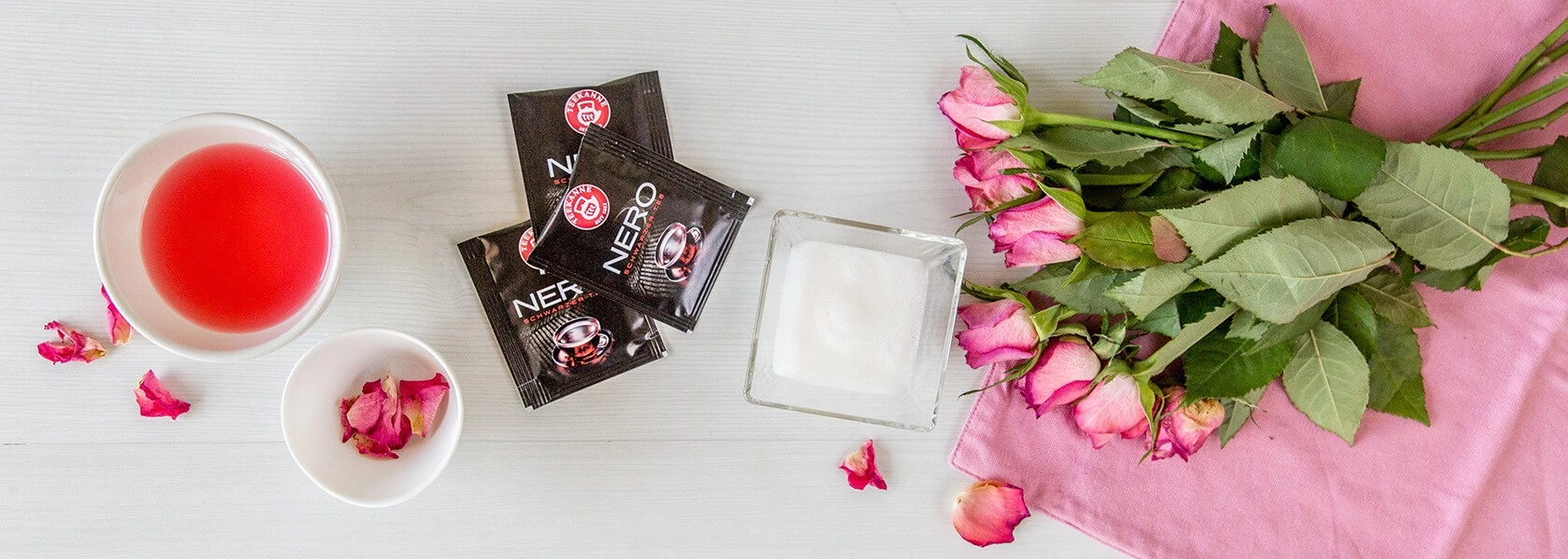 Flach liegend: rotes Getränk, Rosenblätter, Kaffeepäckchen, Zuckerdose, rosa Rosen auf einem rosa Tuch.