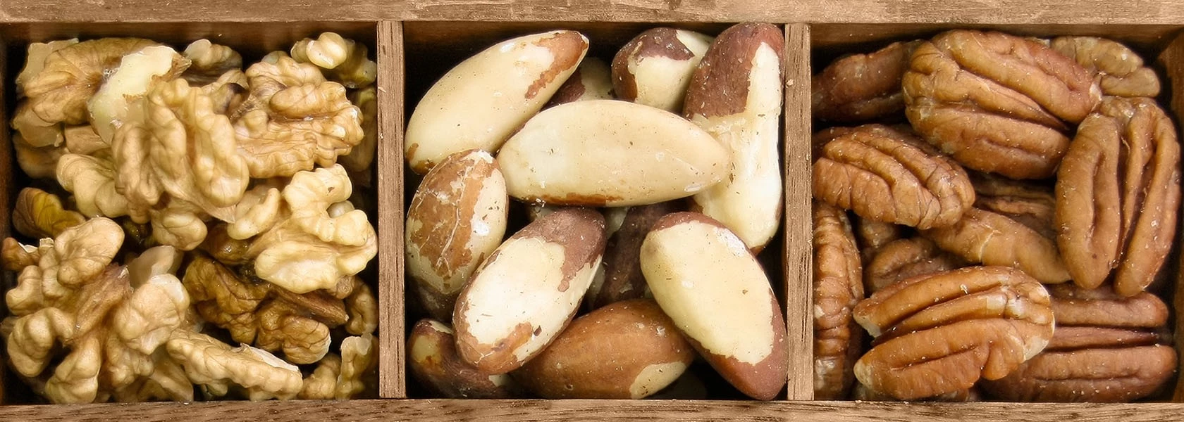Nahaufnahme eines Holztabletts mit Walnüssen, Paranüssen und Pekannüssen in getrennten Abschnitten.