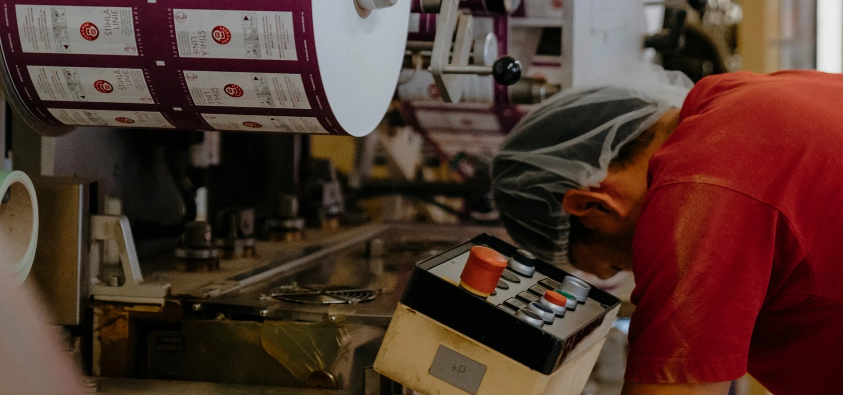 Arbeiter mit Haarnetz bedient Fabrikmaschine; über ihm sind große Rollen mit bedruckten Etiketten zu sehen.