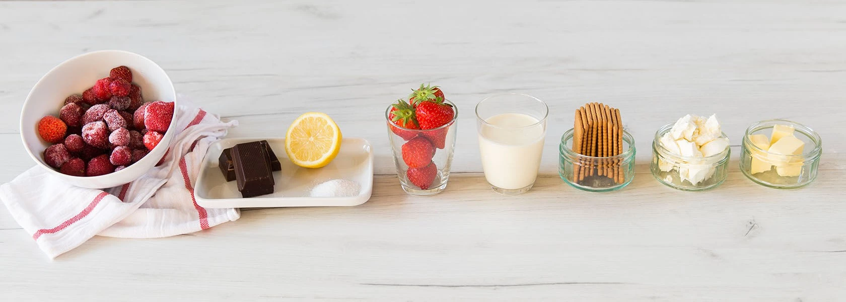 Verschiedene Dessertzutaten in Glasschalen: gefrorene Erdbeeren, Schokolade, Zitrone, Zucker, Milch, Kekse und Schlagsahne.