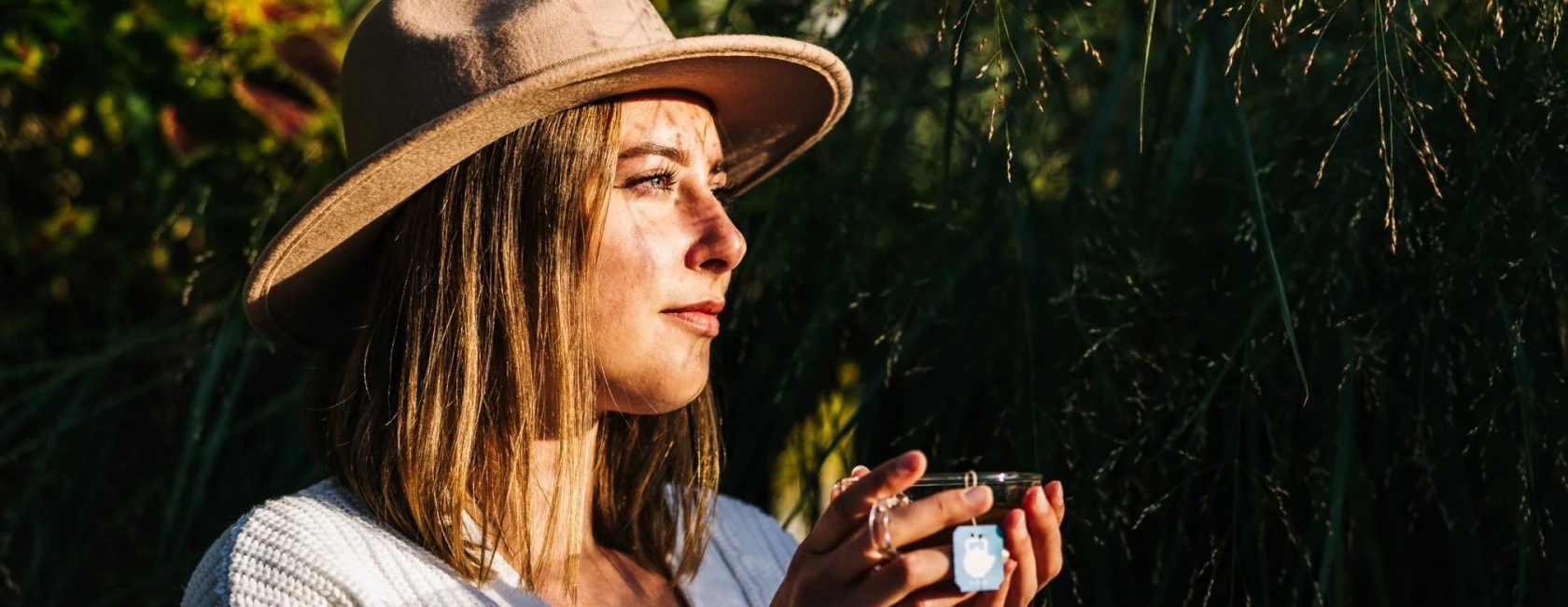 Frau mit breitkrempigem Hut hält eine Tasse und blickt nachdenklich zwischen hohen grünen Pflanzen im Sonnenlicht hindurch.