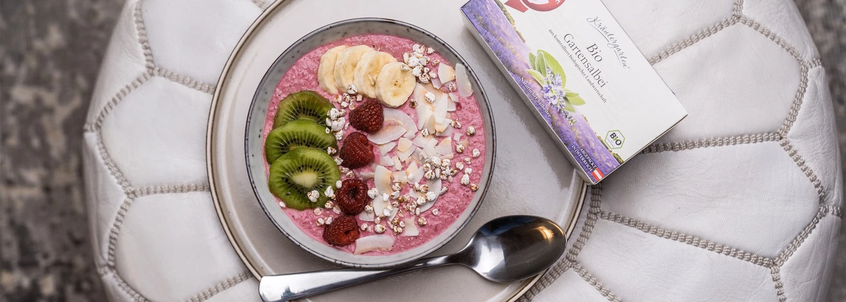 Rosa Smoothie-Schüssel mit Obst, Müsli und Kokosnuss neben einem Löffel und einer Schachtel Bio-Kräutertee.