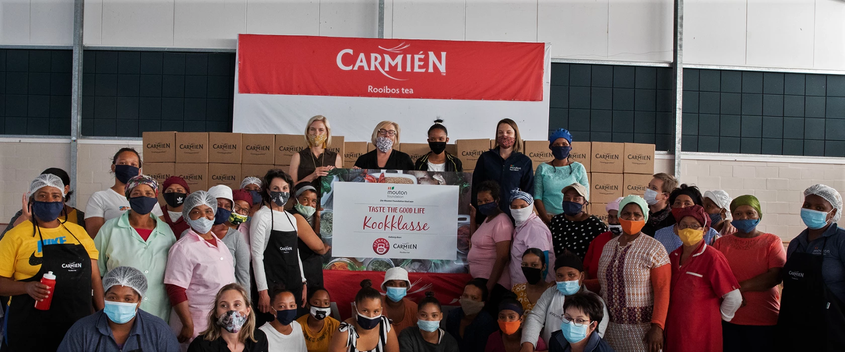 Eine Gruppe mit Masken und Haarnetzen posiert drinnen mit einem „Kookklasse“-Schild und einem Banner mit Carmién-Rooibos-Tee.