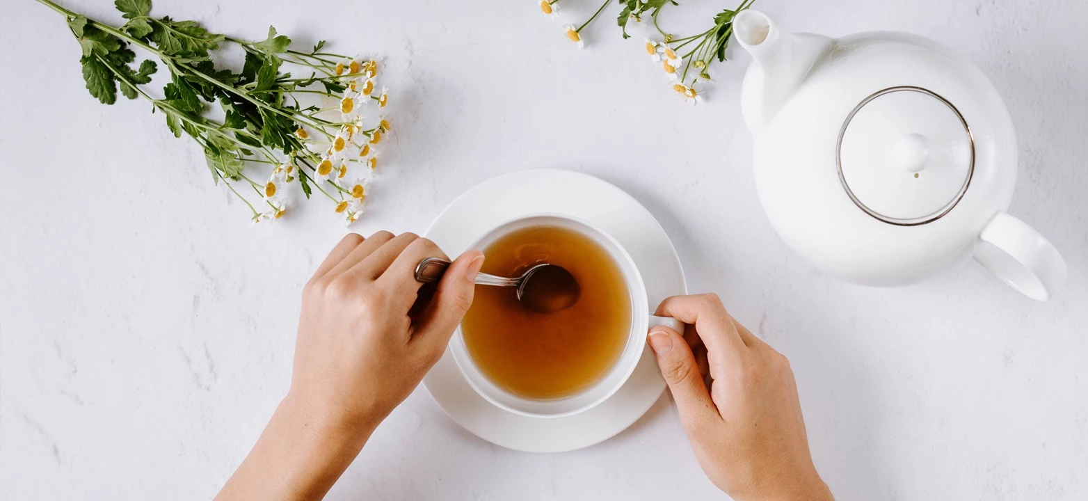 Person rührt Tee in weißer Tasse, mit weißer Teekanne und Kamillenblüten auf heller Oberfläche.