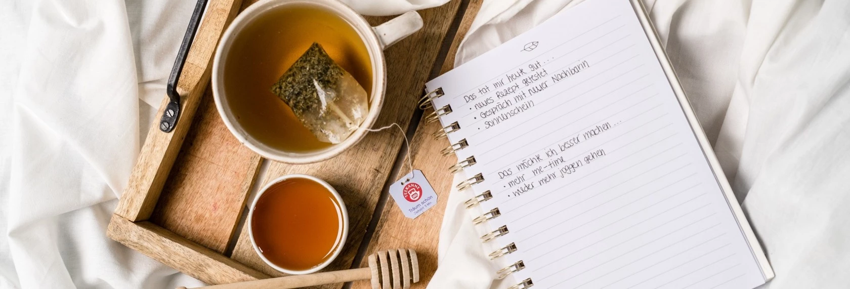 Holztablett mit Tee, Honigschale, Schöpflöffel und offenem Notizbuch auf weißem Stoffhintergrund.