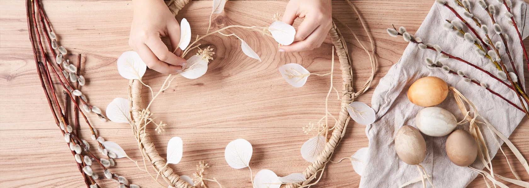 Hände arrangieren einen Kranz aus weißen Blättern auf einem Tisch; daneben liegen Weidenkätzchen und Ostereier.