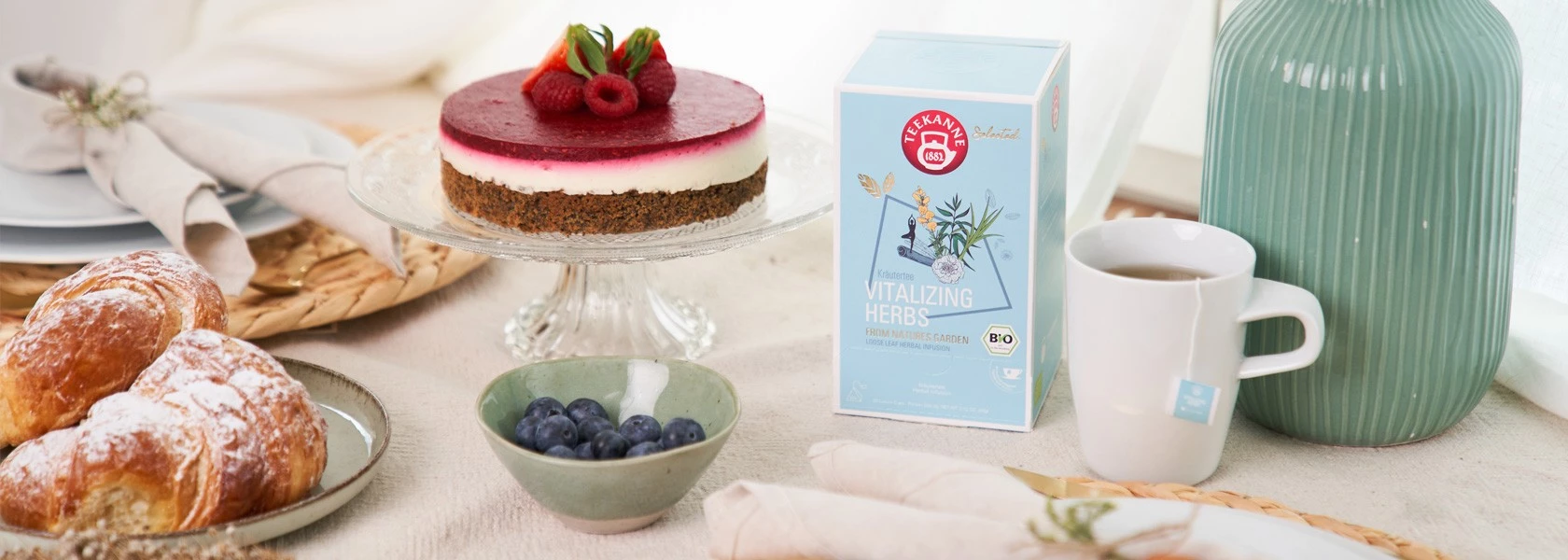 Frühstück mit Croissants, Blaubeeren, Beerenkäsekuchen, Vitalizing Herbs Teebox, Tasse Tee, grüne Vase.
