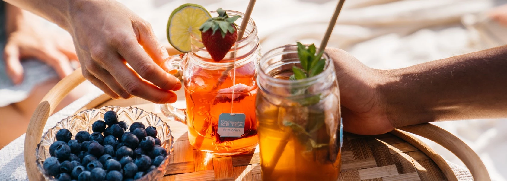 Zwei Hände halten Einmachgläser mit Früchte-Eistee neben Blaubeeren auf einem Holztablett.