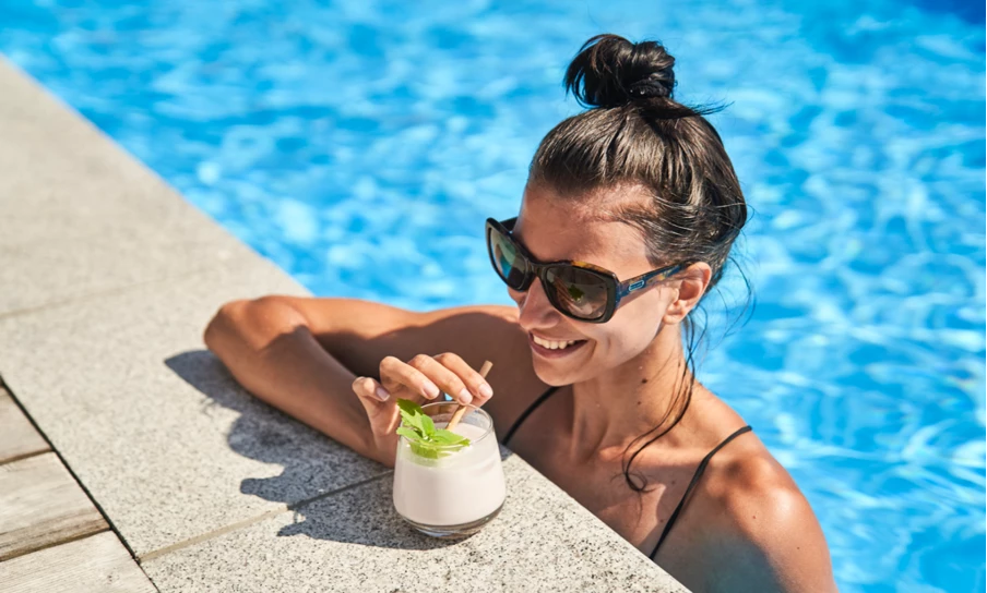 Frau mit Sonnenbrille entspannt sich an einem sonnigen Tag lächelnd im Pool und hält ein cremiges, mit Minze garniertes Getränk in der Hand.
