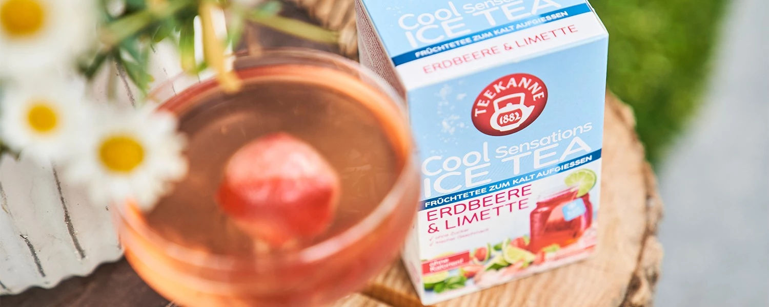 Glas Eistee mit Erdbeere und Teekannenschachtel auf Holz, Gänseblümchen daneben.