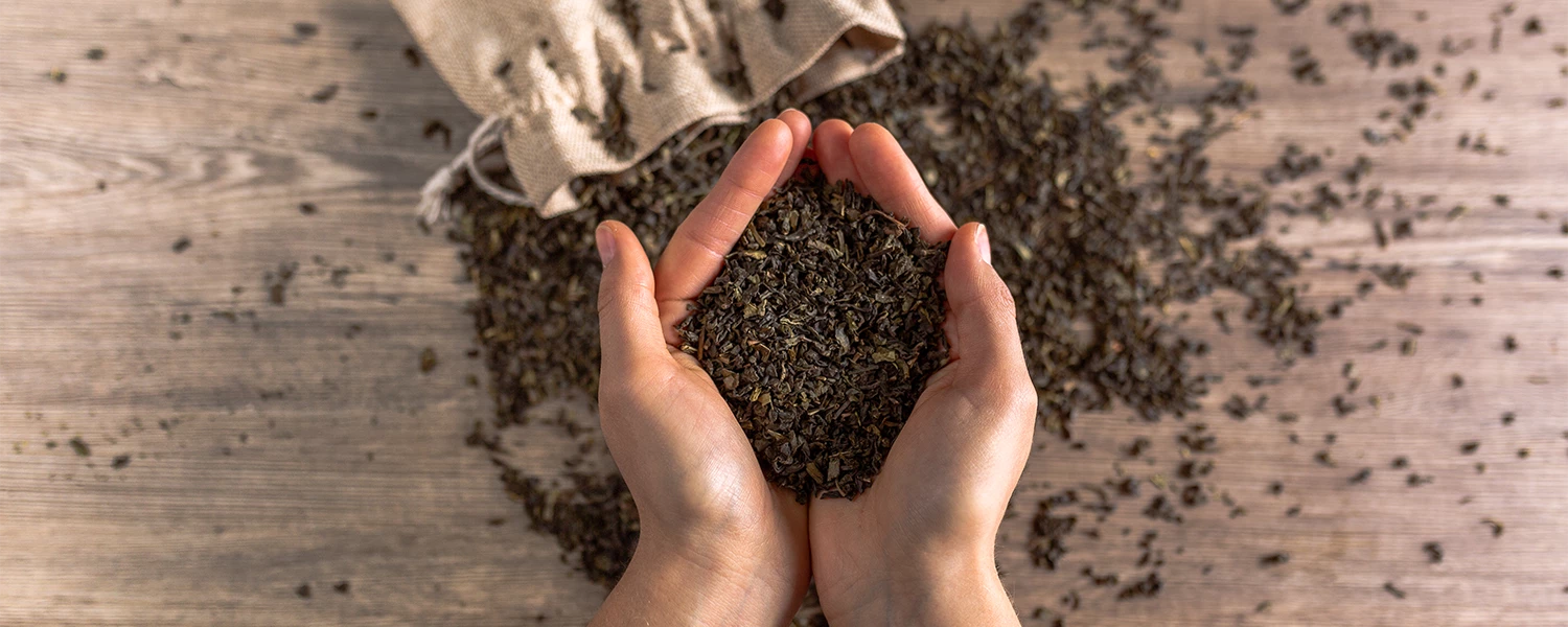 Hände halten lose Teeblätter, weiterer Tee ist auf Holz verstreut und läuft aus einem Leinensack.