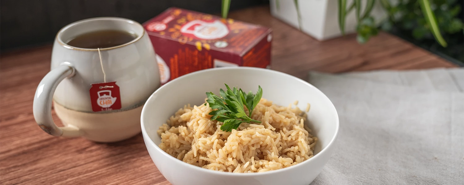 Schüssel Reis mit Petersilie neben einer Tasse Tee, einer Teedose und einer gefalteten Serviette auf dem Tisch.
