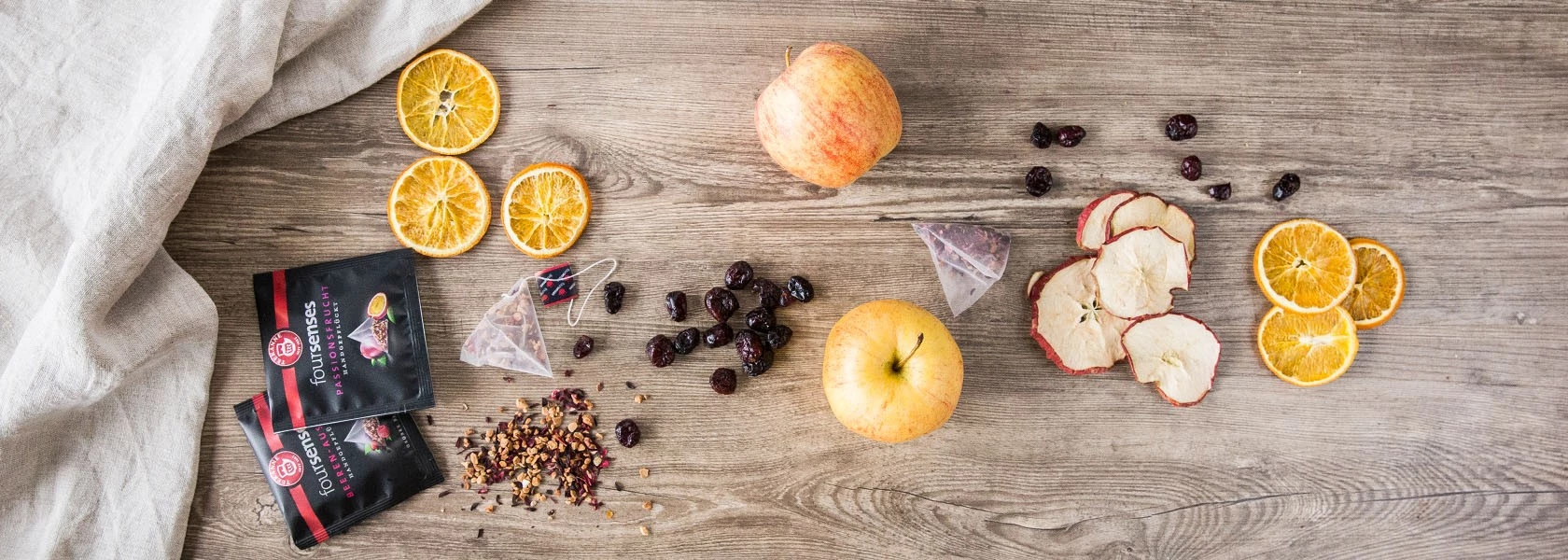 Verschiedene frische Äpfel, Trockenfrüchte und Früchtetee-Päckchen auf Holz angeordnet, daneben liegt ein Leinentuch.