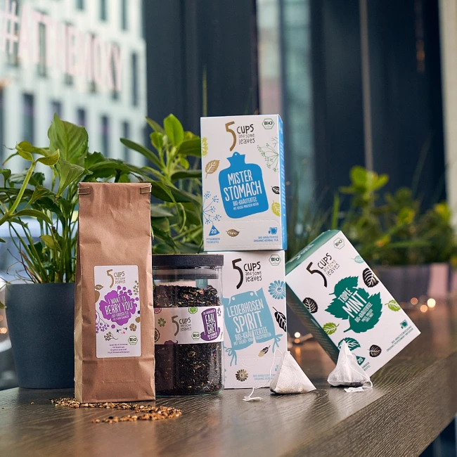 5 Tassen und einige Blätter Teeprodukte auf einem Holztisch mit Topfpflanzen, im Hintergrund Stadtansicht.