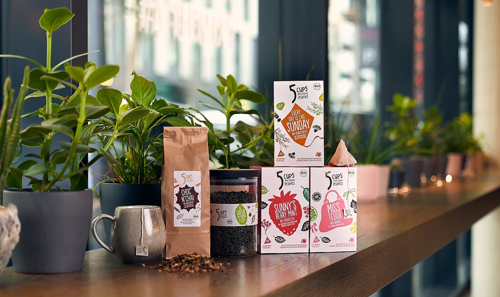 Verschiedene Teeprodukte und eine Tasse stehen auf der Fensterbank mit grünen Pflanzen; vor dem Fenster sind Stadtgebäude zu sehen.