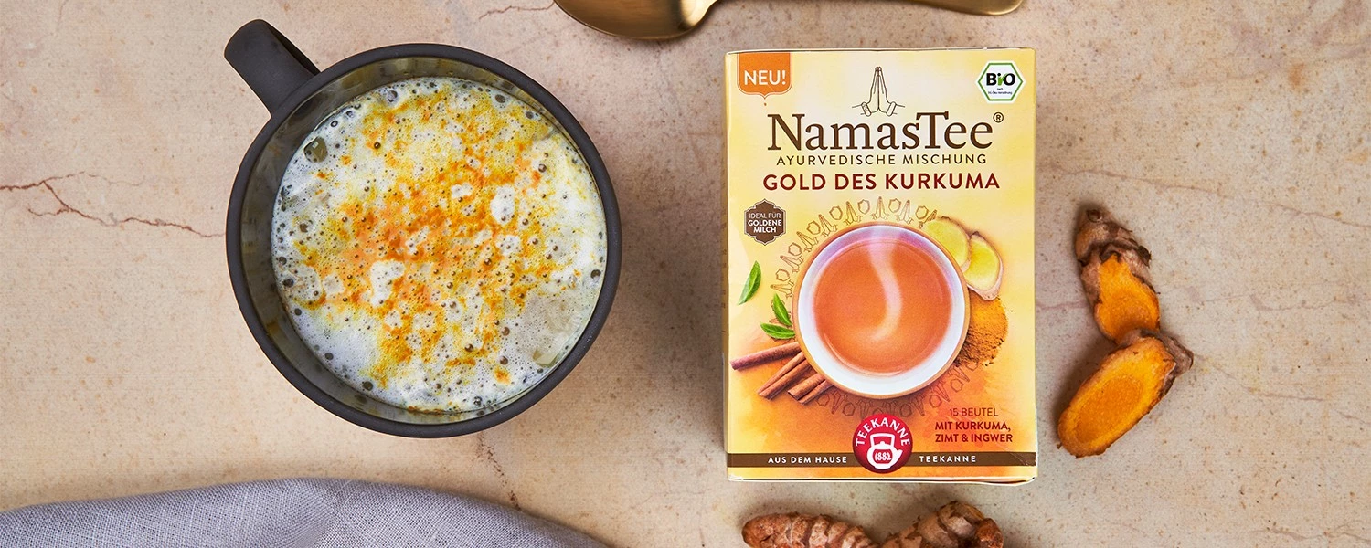 Kurkuma-Latte in einer Tasse neben einer goldenen Teedose von NamasTee, Kurkumawurzeln und einem goldenen Löffel.