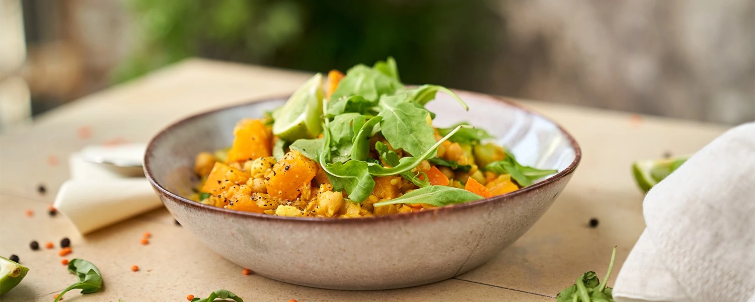 Bunter Salat mit Kürbis, Kichererbsen, Rucola in einer Keramikschüssel; daneben Limettenspalten und Blattgemüse.