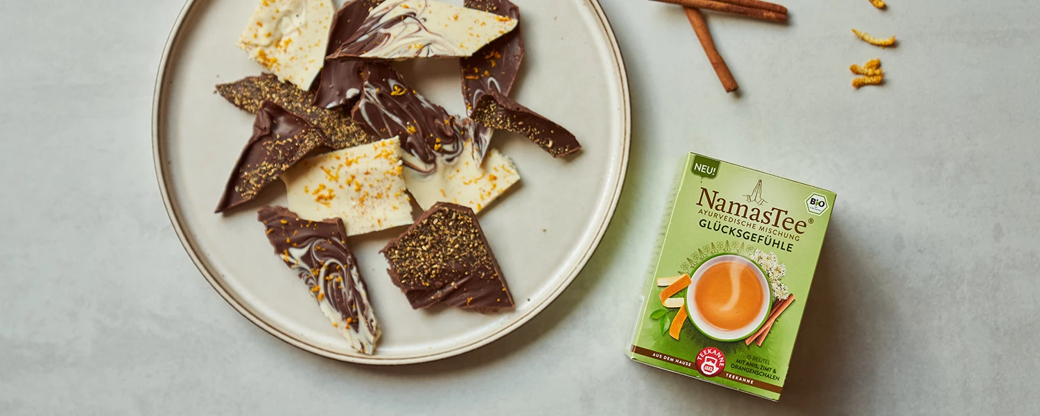 Schokoladenrinde mit Gewürzen neben einer Schachtel NamasTee-Tee, Zimtstangen und Teeblättern.