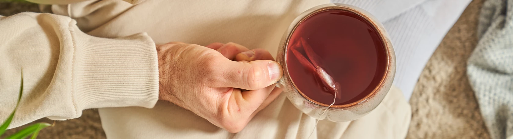 Person im beigen Pullover hält eine Glastasse mit rotem Tee, Teebeutelschnur sichtbar, gemütliche Umgebung.