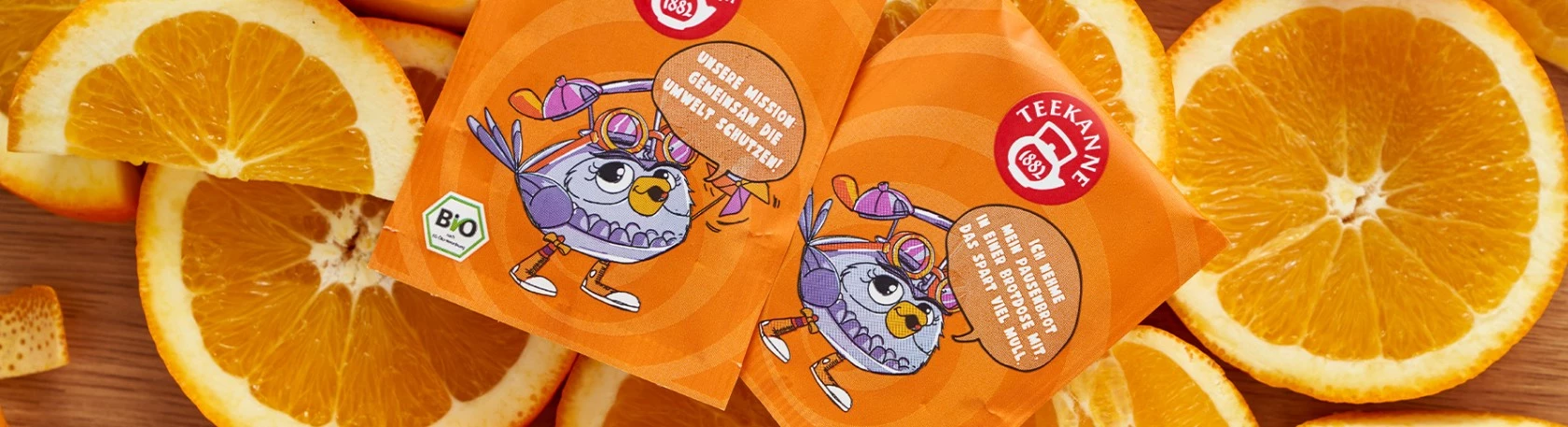 Zwei orangefarbene Teekanne Bio-Teebeutel mit Cartoon-Vogel, umgeben von frischen Orangenscheiben.
