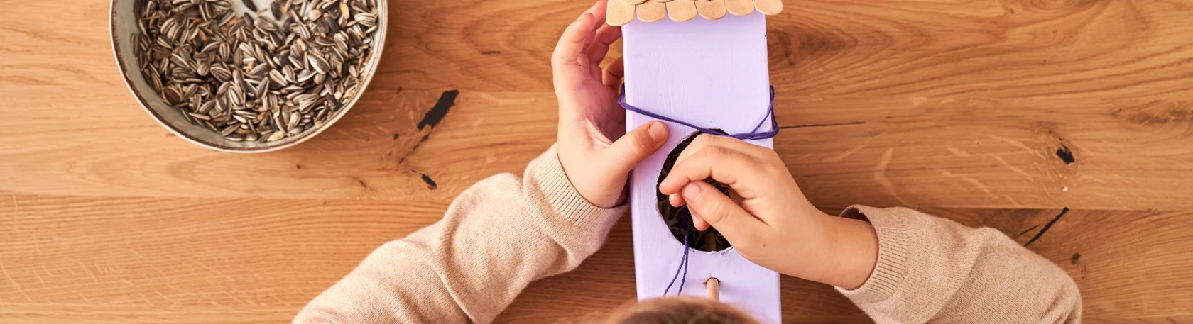 Kind legt Samen in ein selbstgemachtes lila Vogelhäuschen, Schüssel mit Sonnenblumenkernen steht in der Nähe auf einem Holztisch.