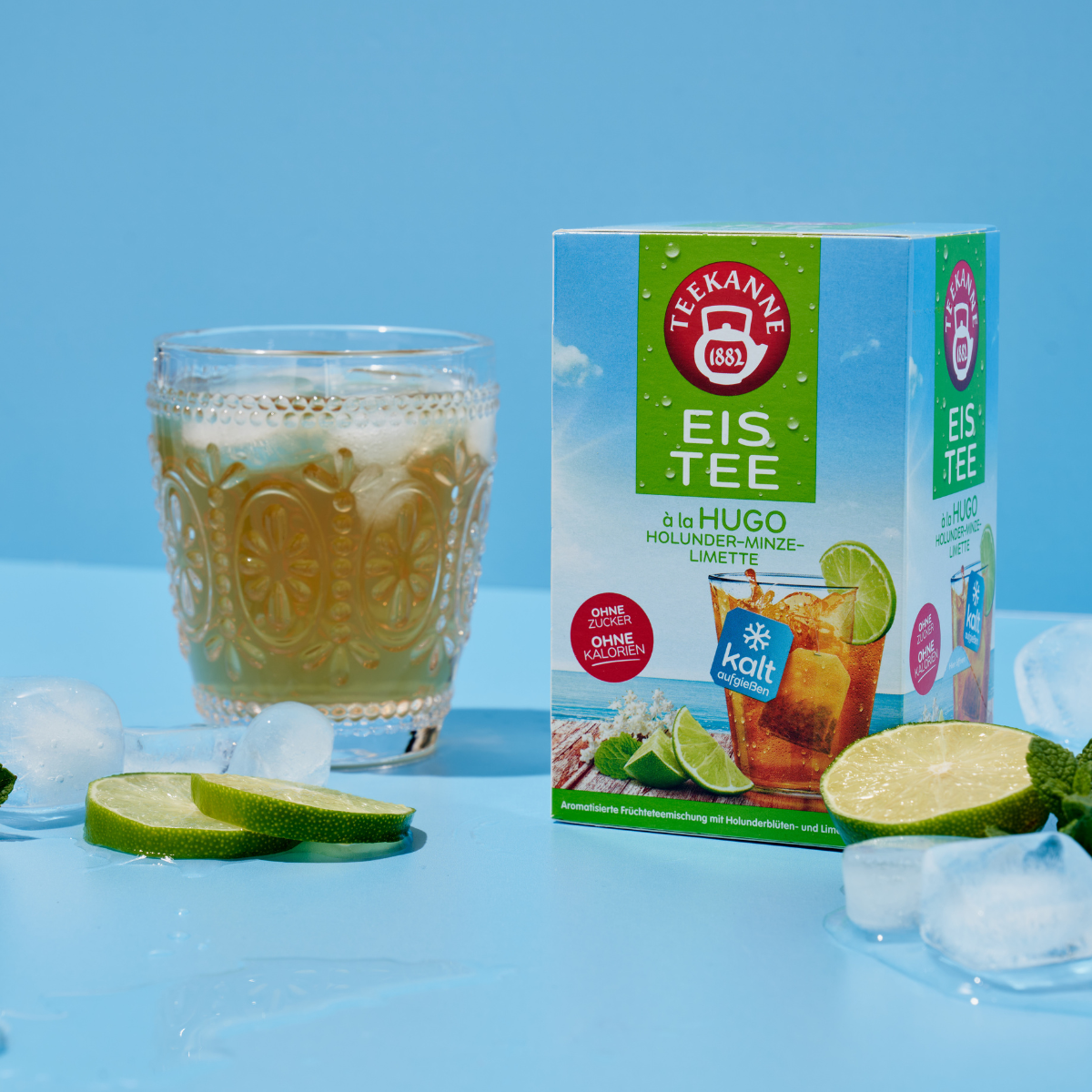 Eine Schachtel Tee à la Hugo steht auf einer blauen Fläche neben einem Glas Eistee, Eiswürfeln und Limettenscheiben vor einem hellblauen Hintergrund.