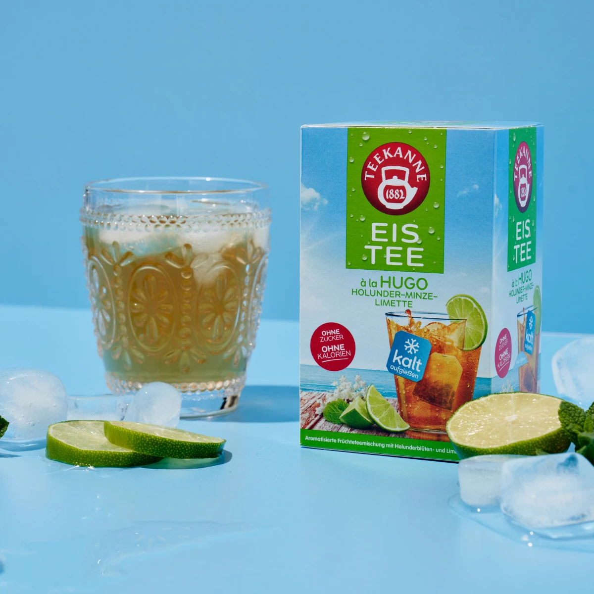 Eine Schachtel Tee à la Hugo steht auf einer blauen Fläche neben einem Glas Eistee, Eiswürfeln und Limettenscheiben vor einem hellblauen Hintergrund.