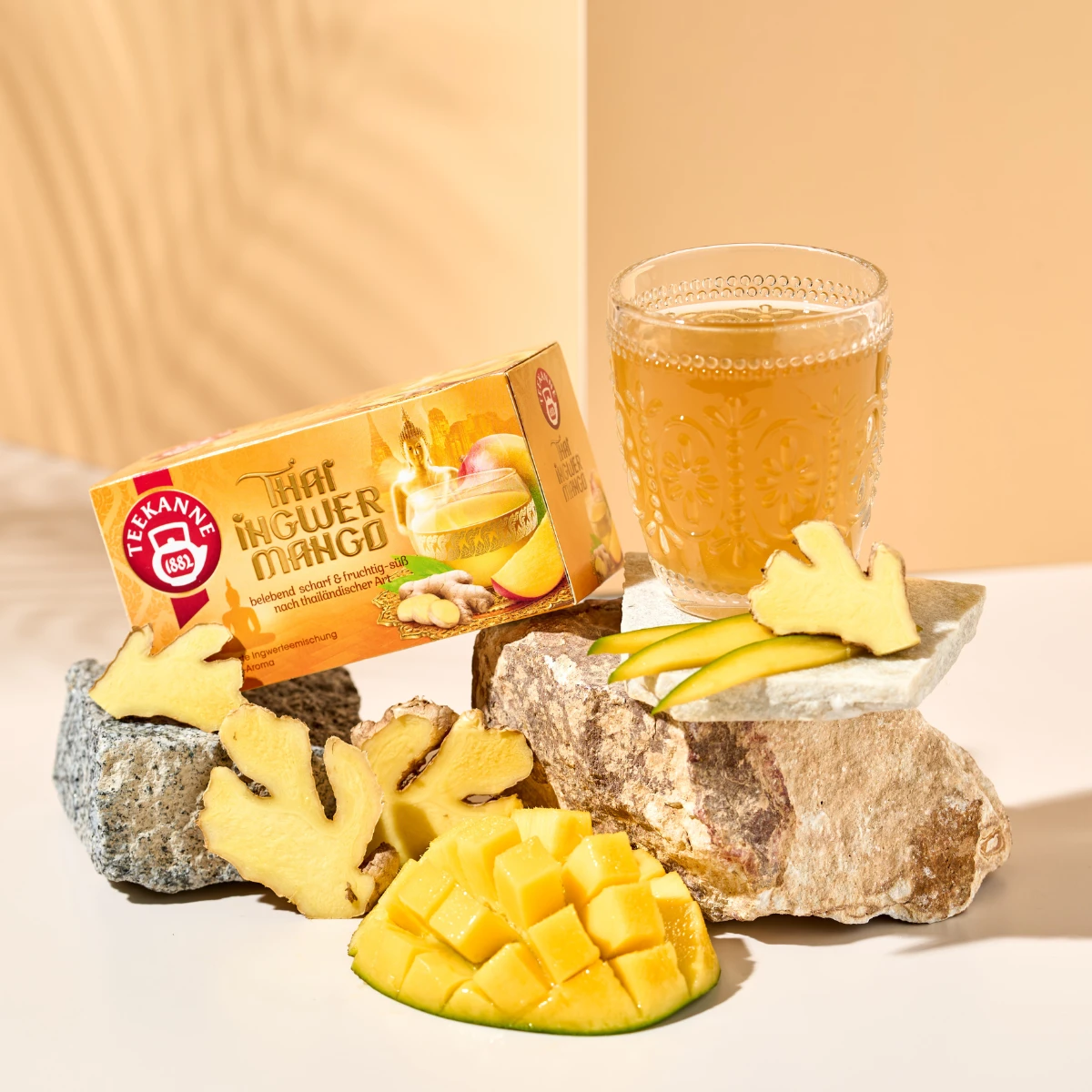 Ein Glas Thai Ingwer Mango-Tee steht neben einer Schachtel mit der Aufschrift „Thai Ingwer Mango“ mit frischen Mangoscheiben und Ingwerstücken, die unter warmem, hellem Licht auf Steinen angeordnet sind.