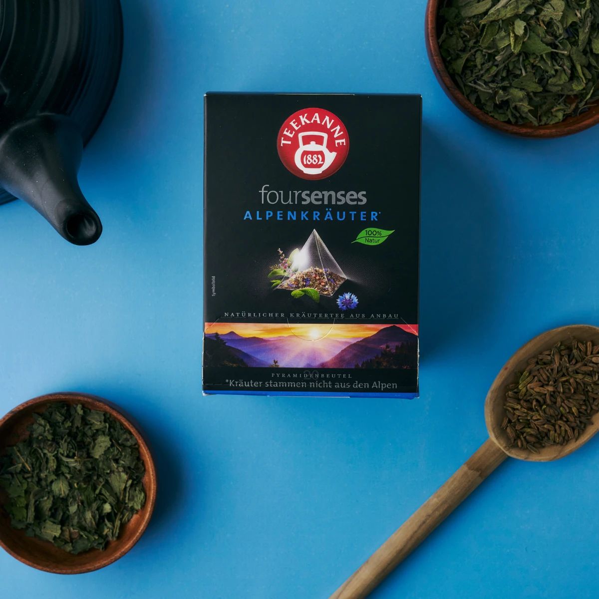 Eine Schachtel Alpenkräuter-Tee steht auf einer blauen Fläche, umgeben von frischen getrockneten Kräutern in Schalen und einer Holzschaufel. In der Nähe ist ein Teil einer schwarzen Teekanne zu sehen.