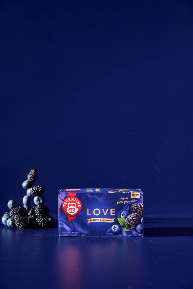 Eine Schachtel LOVE Blaubeer-Brombeer-Tee steht auf einer blauen Fläche, hinter der sich frische Blaubeeren und Brombeeren stapeln, vor einem tiefblauen Hintergrund.