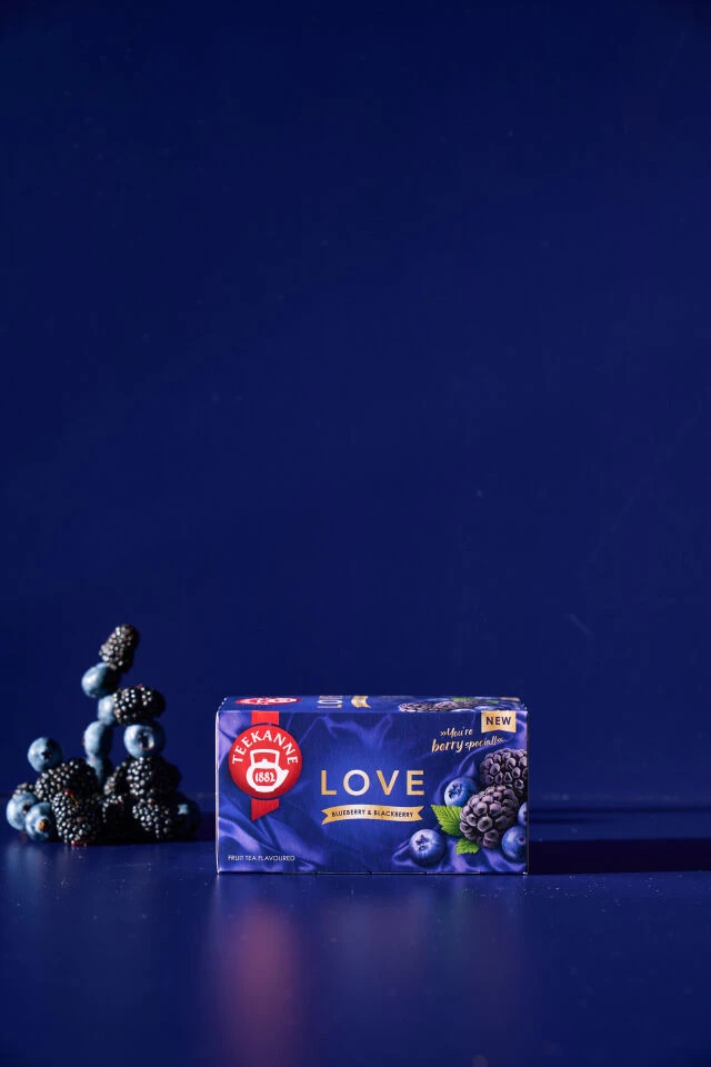 Eine Schachtel LOVE Blaubeer-Brombeer-Tee steht auf einer blauen Fläche, hinter der sich frische Blaubeeren und Brombeeren stapeln, vor einem tiefblauen Hintergrund.