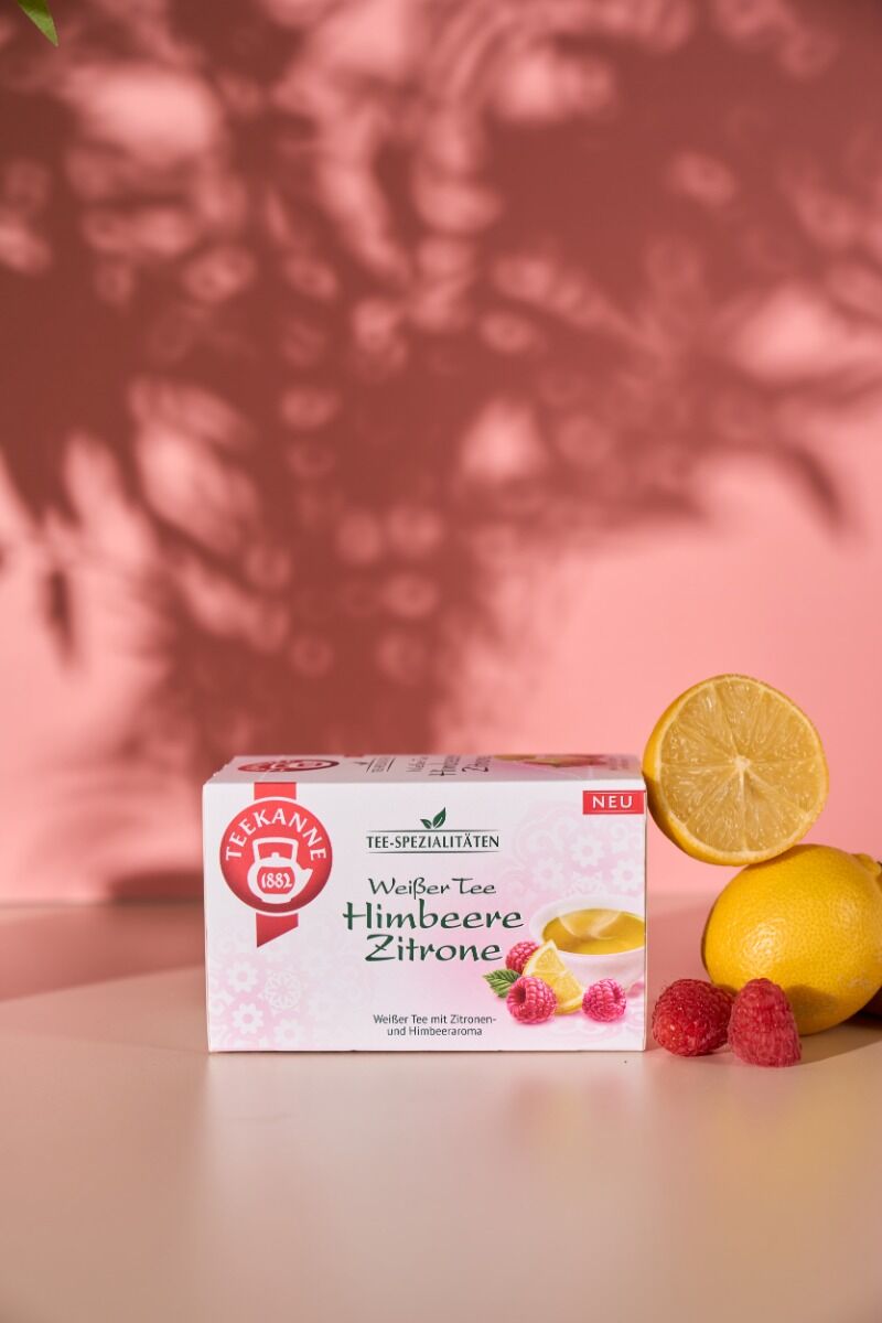 Eine Schachtel Weißer Tee Himbeere Zitrone steht vor einem rosafarbenen Hintergrund, neben einer ganzen Zitrone, einer Zitronenhälfte und Himbeeren, mit Blattschatten an der Wand.